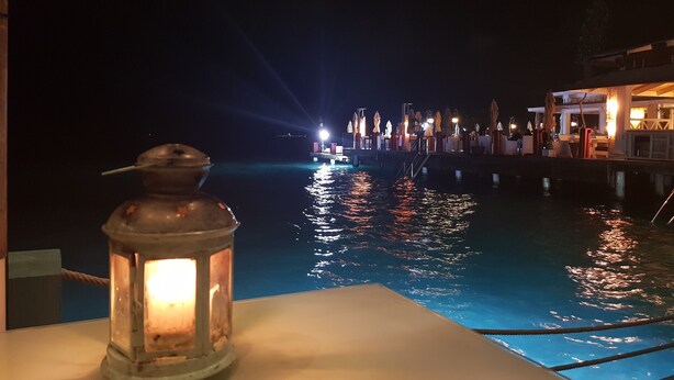 A night scene with a calm body of water reflecting lights, a lantern on a table, and a dock with umbrellas and a building in the background. - KSL Residence (Boca Chica)