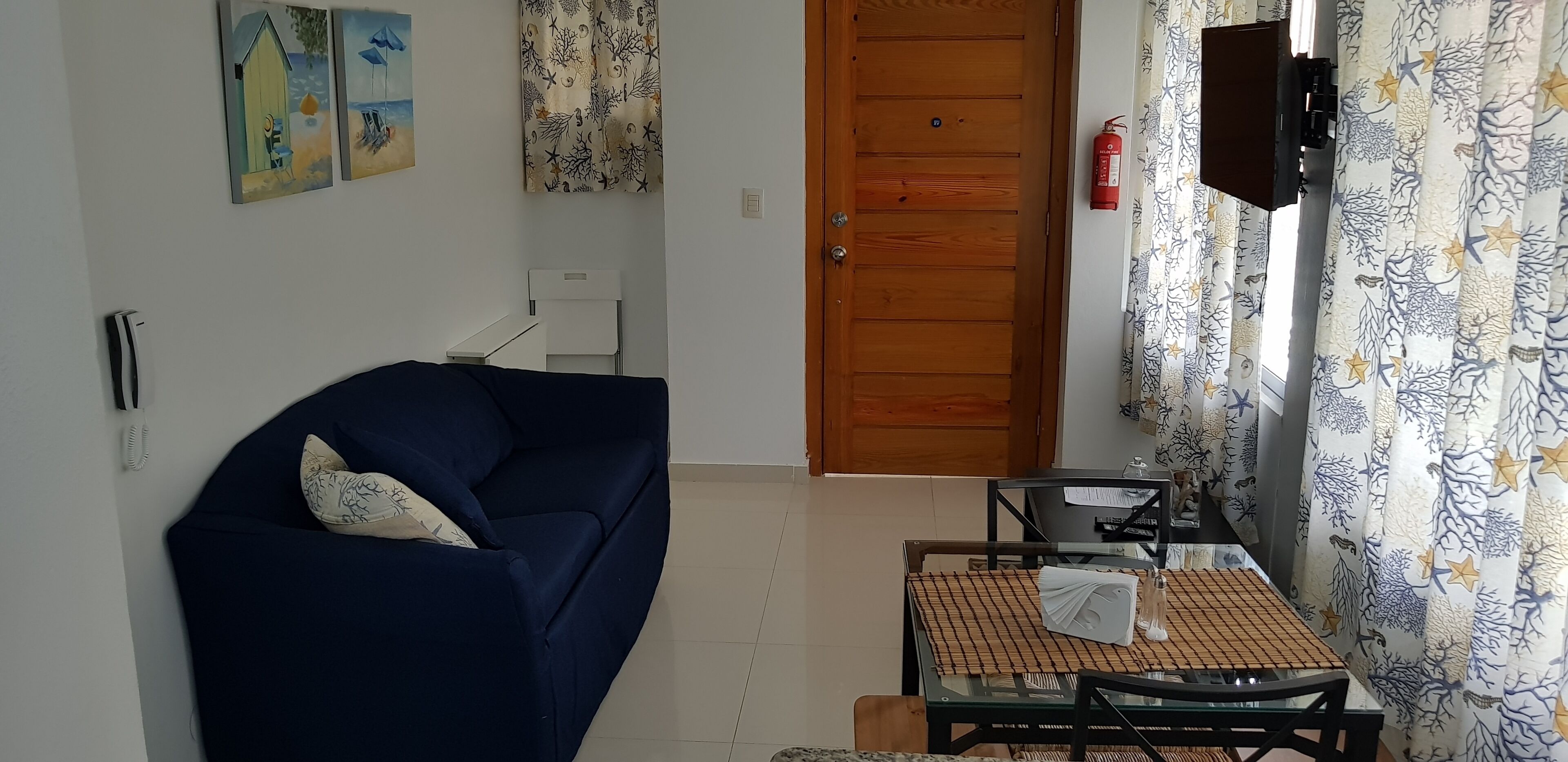 Un salón con un sofá azul oscuro, una mesa de vidrio con camino de mesa a cuadros, y una puerta de madera.