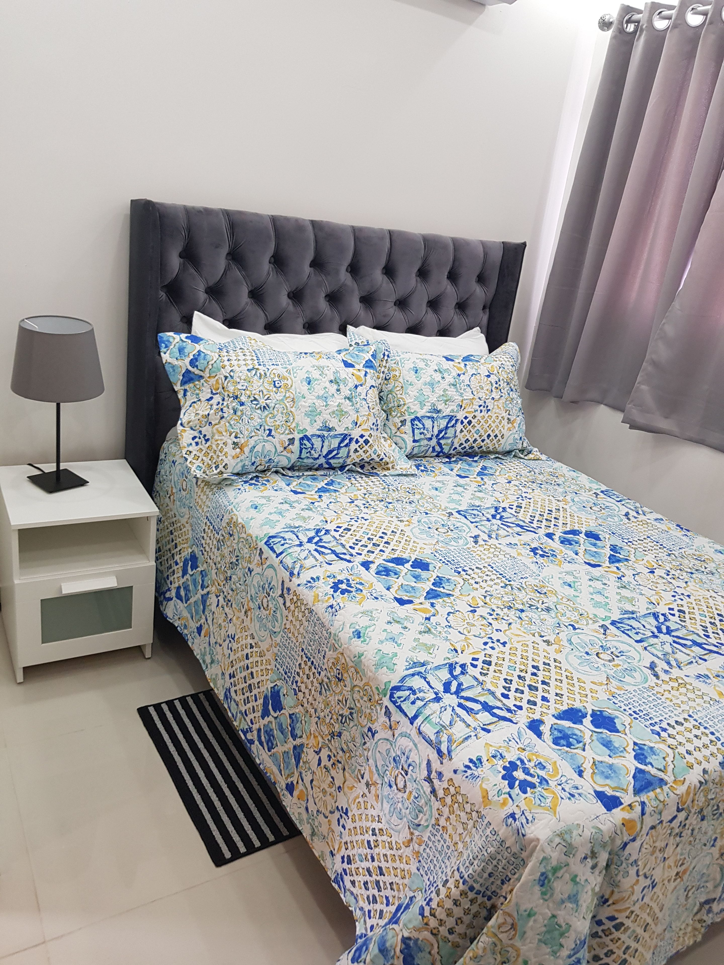 A neatly made bed with a patterned bedspread, a bedside lamp, and a nightstand with a drawer.