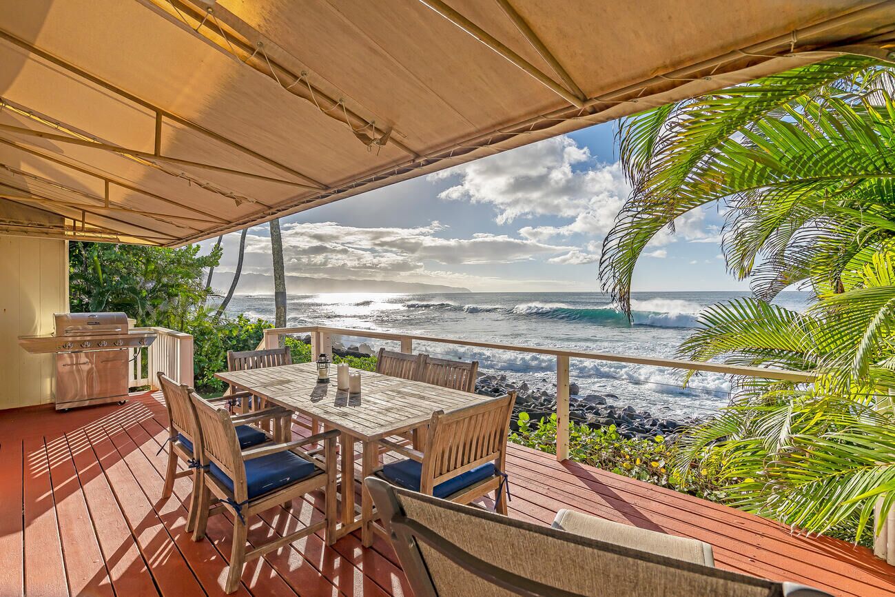 A wooden deck with a table and chairs, overlooking a beach with waves and a cloudy sky.