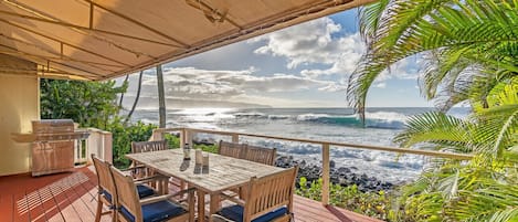A wooden deck with a table and chairs, overlooking a beach with waves and a cloudy sky.
