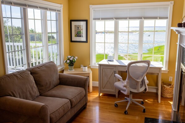 A room with a brown sofa, a desk with a computer, and a view of a body of water through large windows. - Ocean Views From Every Room, Beach 30 Seconds Away! (West Hyannisport)