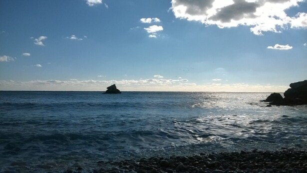 Paisaje costero con un litoral rocoso, mar tranquilo y una formación rocosa distante.