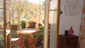 Un balcón abierto con una mesa y sillas de madera, plantas en macetas y un pequeño armario de madera.
