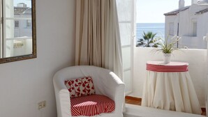 Un sillón blanco con almohadones a rayas rojas y blancas, una mesita redonda con una planta en maceta y vista al mar a través de una ventana con cortinas color beige.