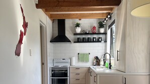 Una cocina compacta con armarios blancos, una estufa y una campana extractora negra. Hay un cubo de basura pequeño y un banco en la habitación.