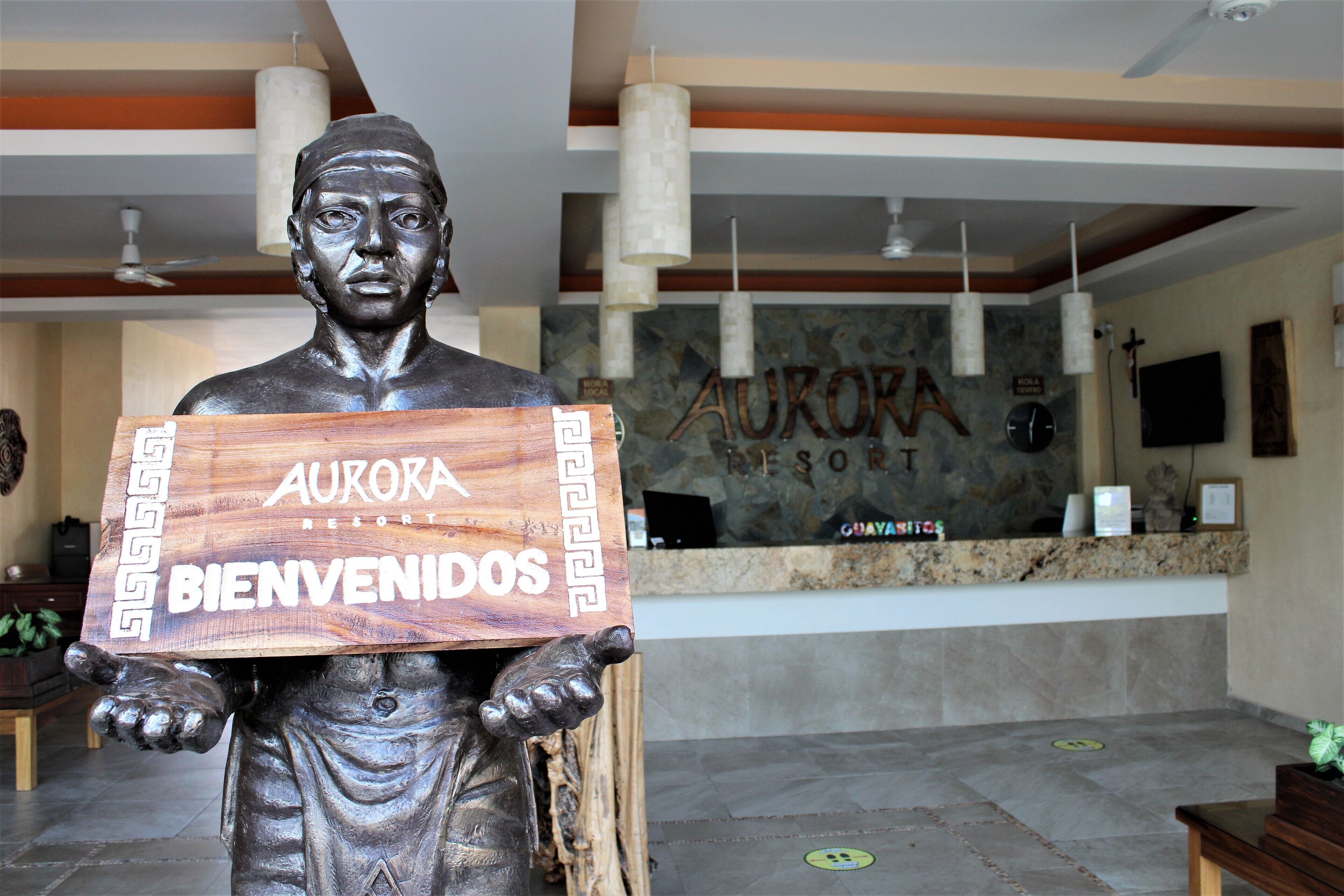 Eine bronzene Statue, die ein Holzschild mit der Aufschrift "AURORA RESORT BIENVENIDOS" hält, in einer Empfangshalle mit Empfangsschalter, Steinwänden und hängenden Pendelleuchten.