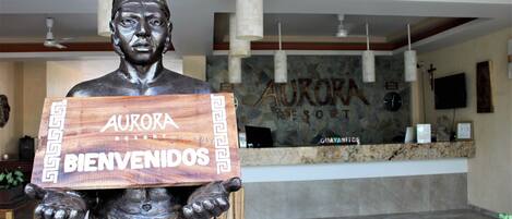 Eine bronzene Statue, die ein Holzschild mit der Aufschrift "AURORA RESORT BIENVENIDOS" hält, in einer Empfangshalle mit Empfangsschalter, Steinwänden und hängenden Pendelleuchten.