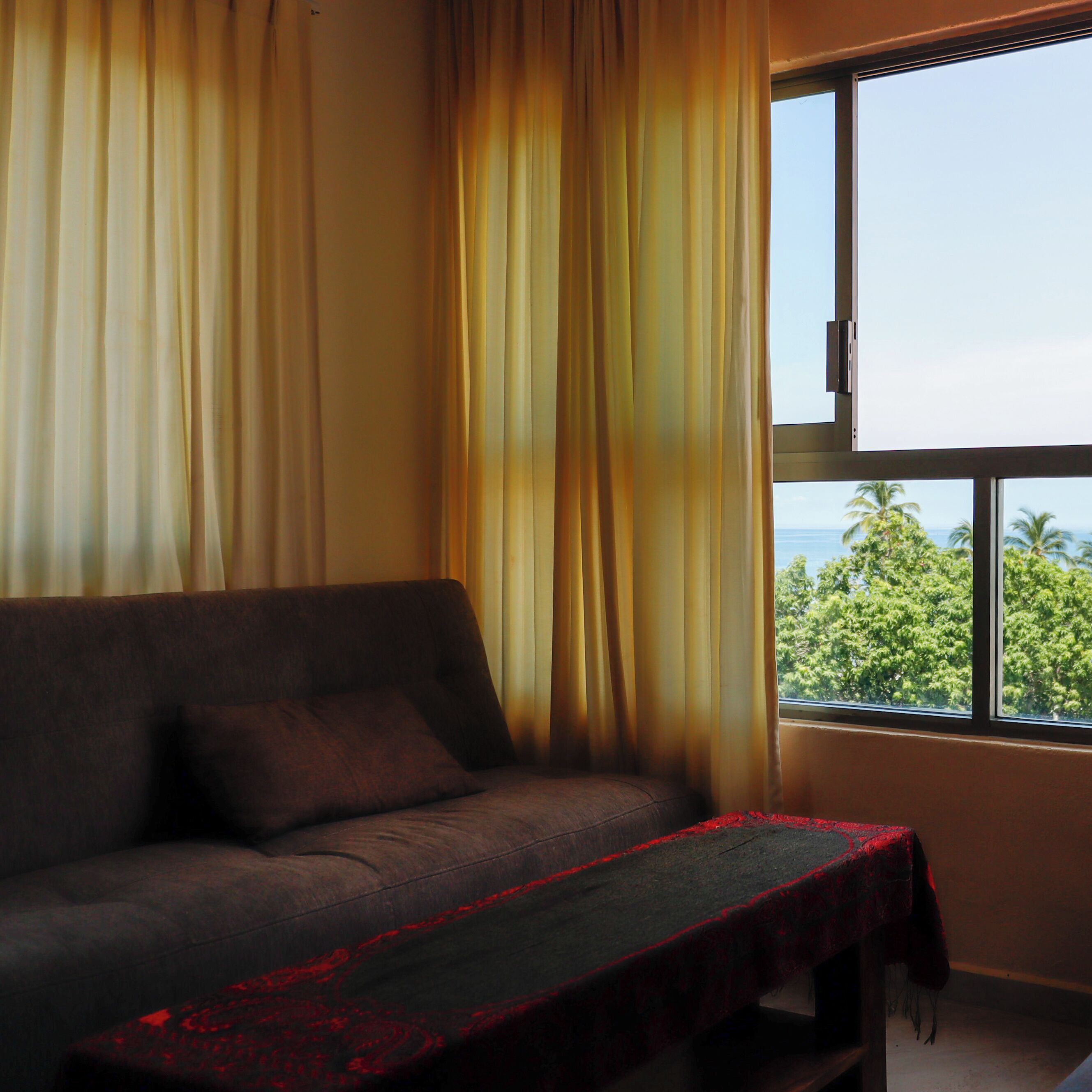 Ein Zimmer mit einer Couch, einem Fenster mit Blick auf Bäume und einem Tisch mit rotem Tischläufer.