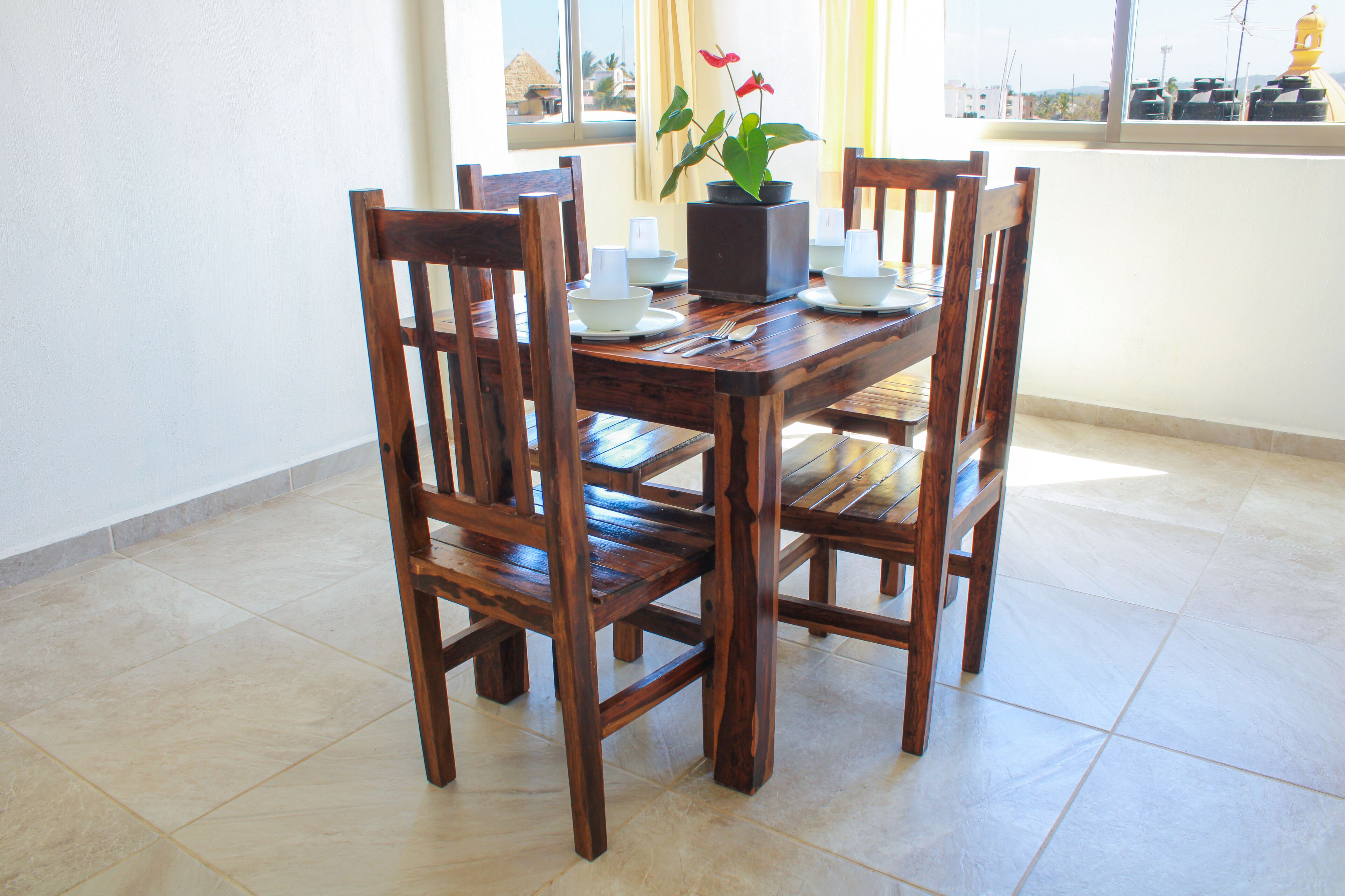 A wooden dining table with four chairs, a potted plant, and white plates and cutlery.