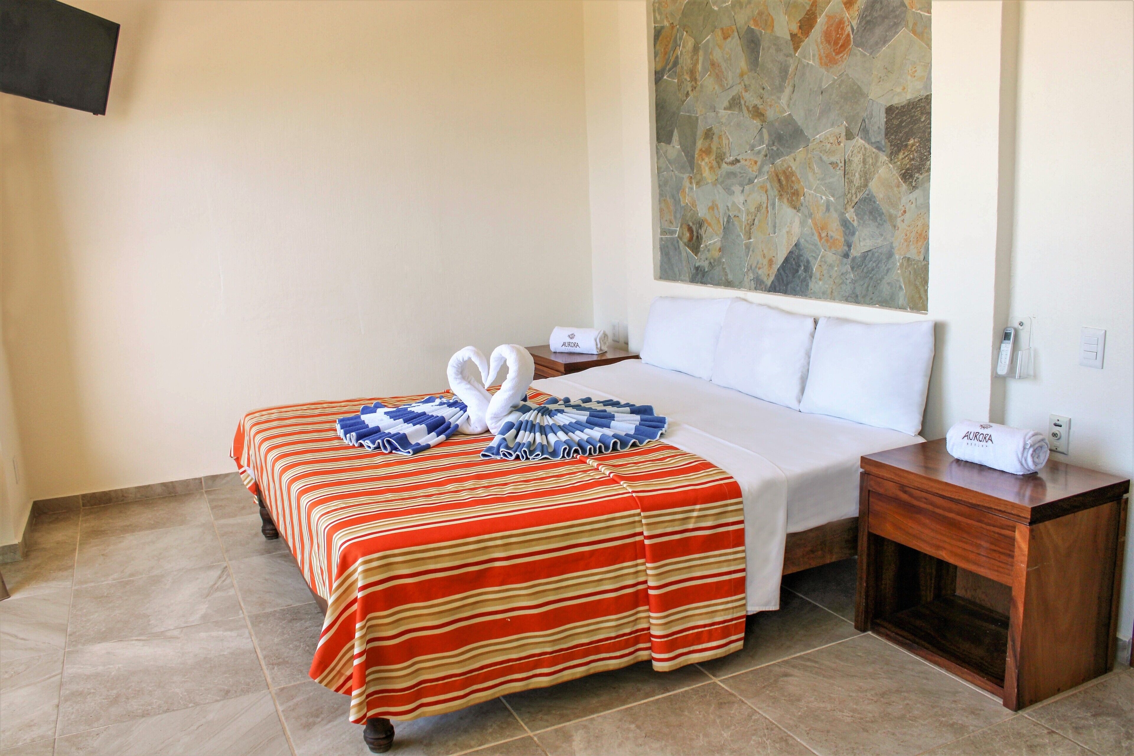 Habitación de hotel con cama, mesita de noche, televisor montado en la pared y pared con diseño de piedra.