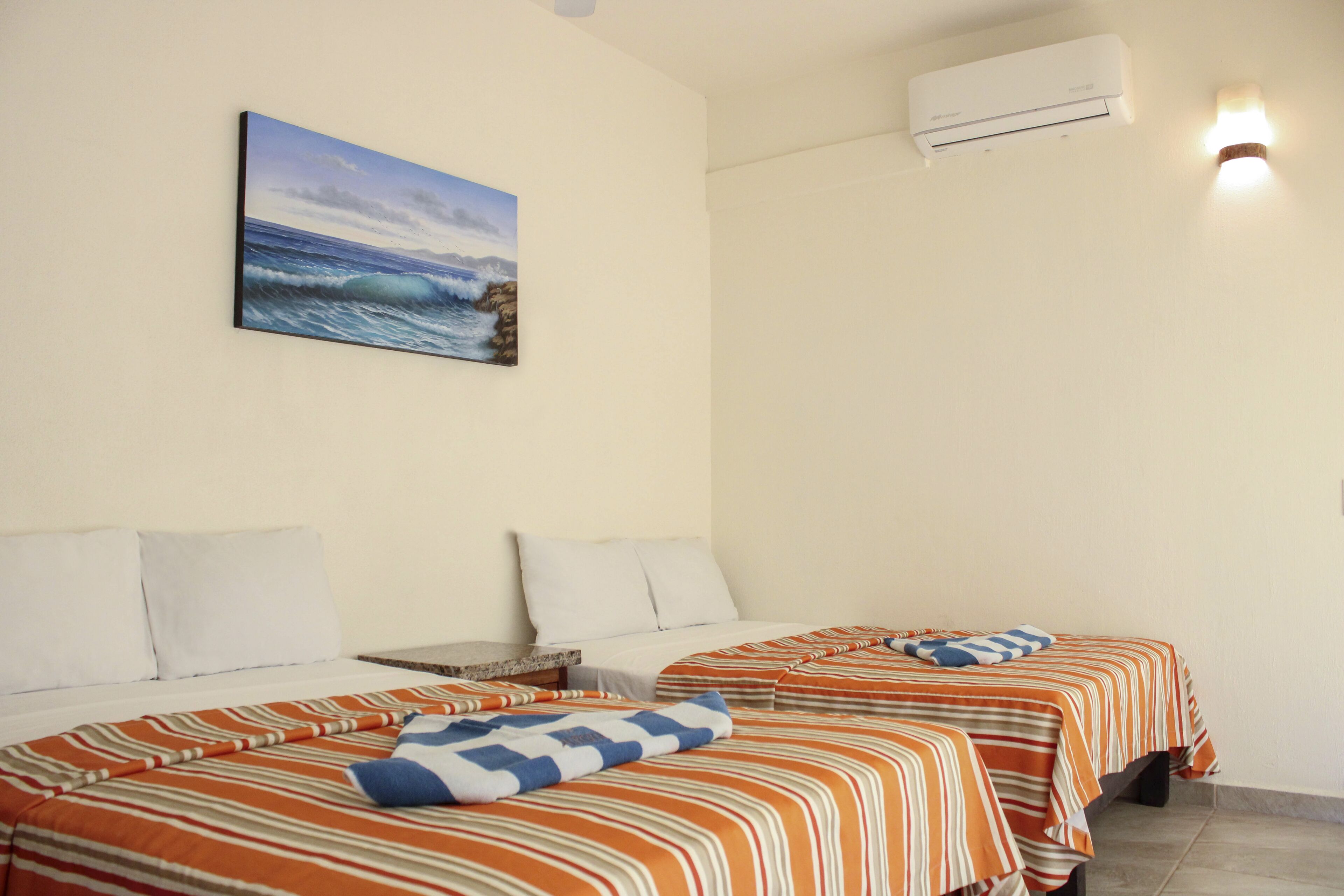 A hotel room with two beds, each with striped bedding and pillows, a painting of a beach scene, and an air conditioning unit on the wall.