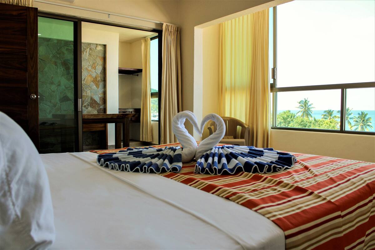 A hotel room with a bed, a view of palm trees, and a window with curtains.