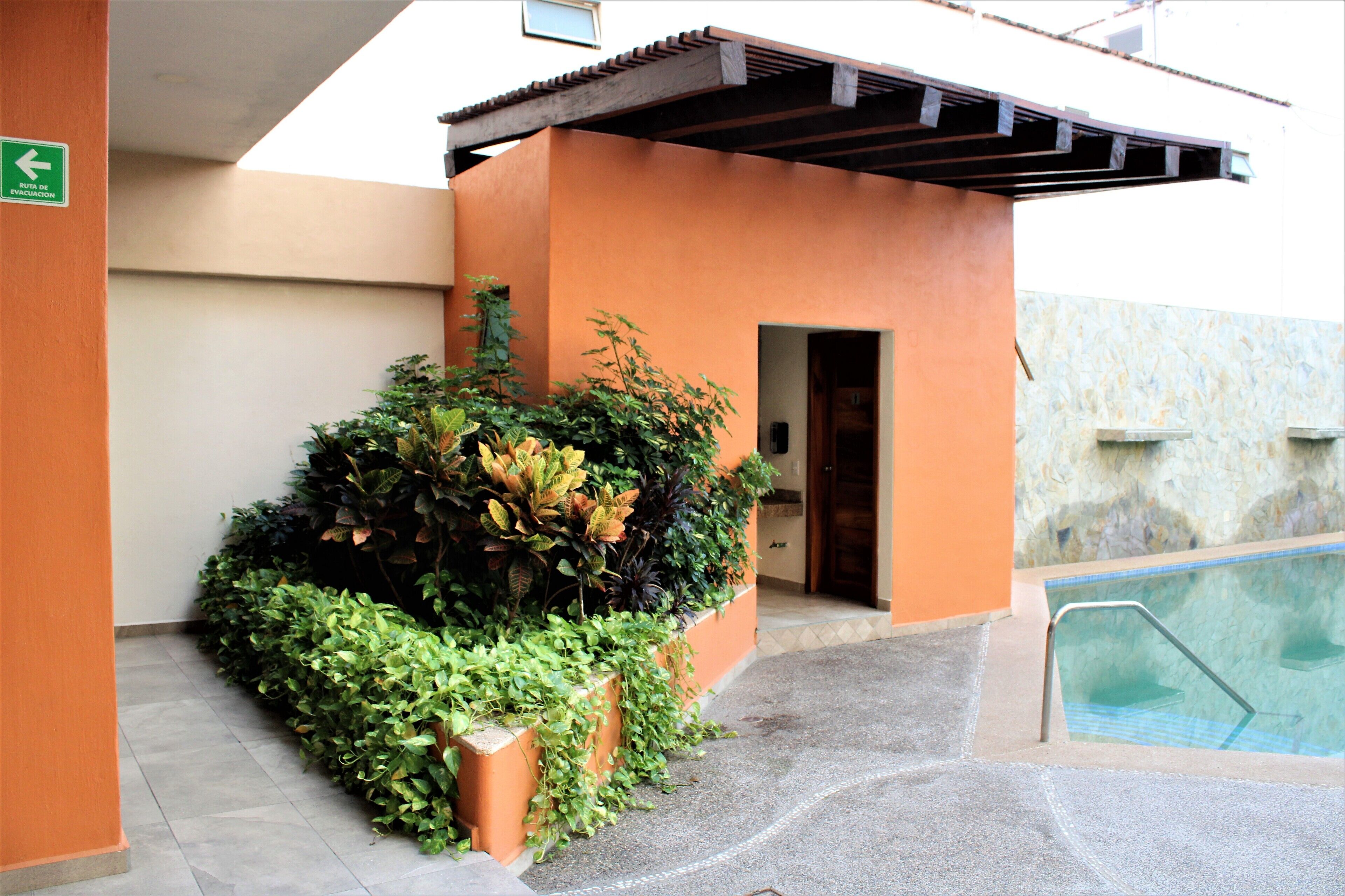 An outdoor pool area with a concrete pathway leading to a door, surrounded by lush green plants and an orange building facade.