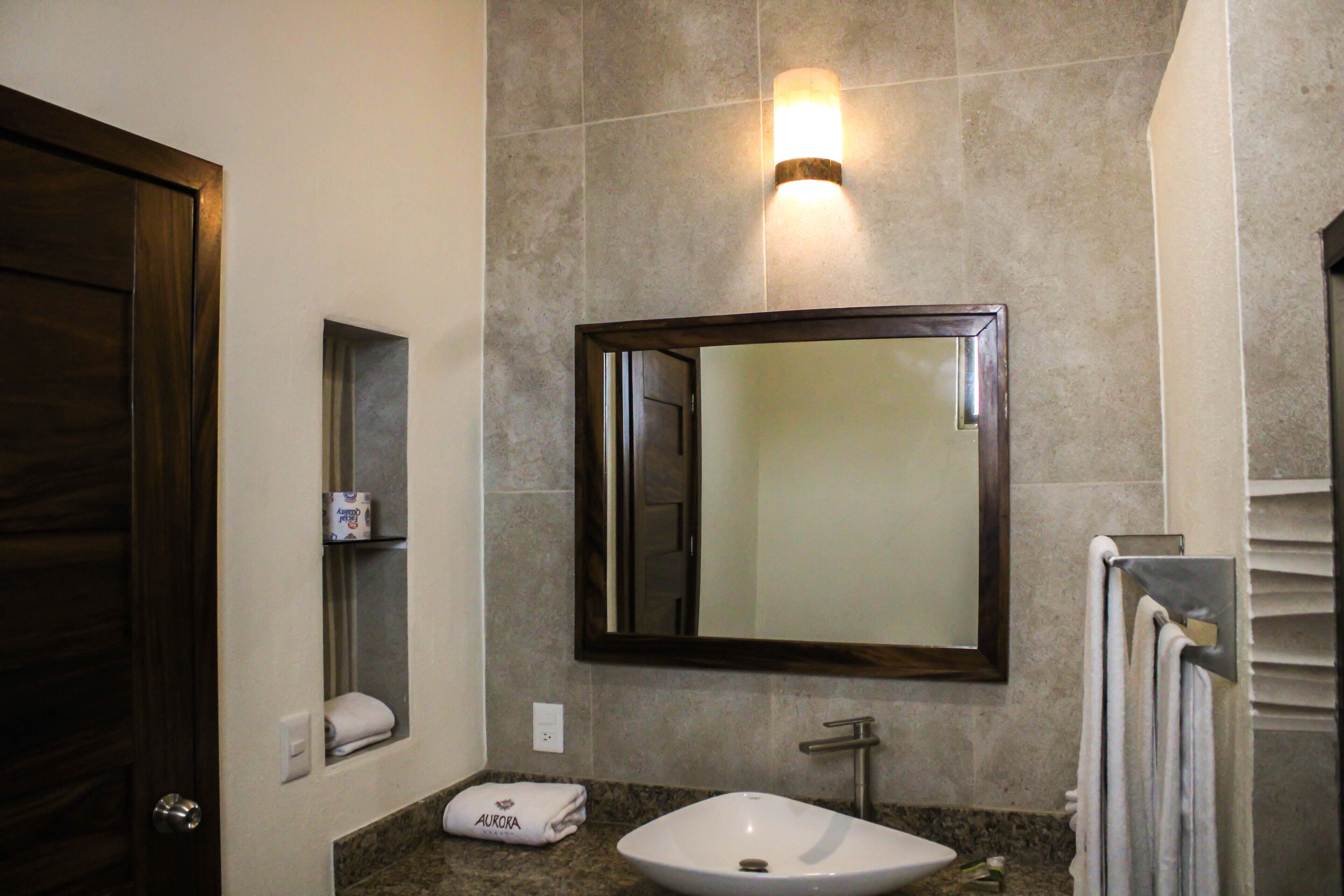 A bathroom with a marble countertop, a white sink, a large mirror, a towel rack, and a wall-mounted light.