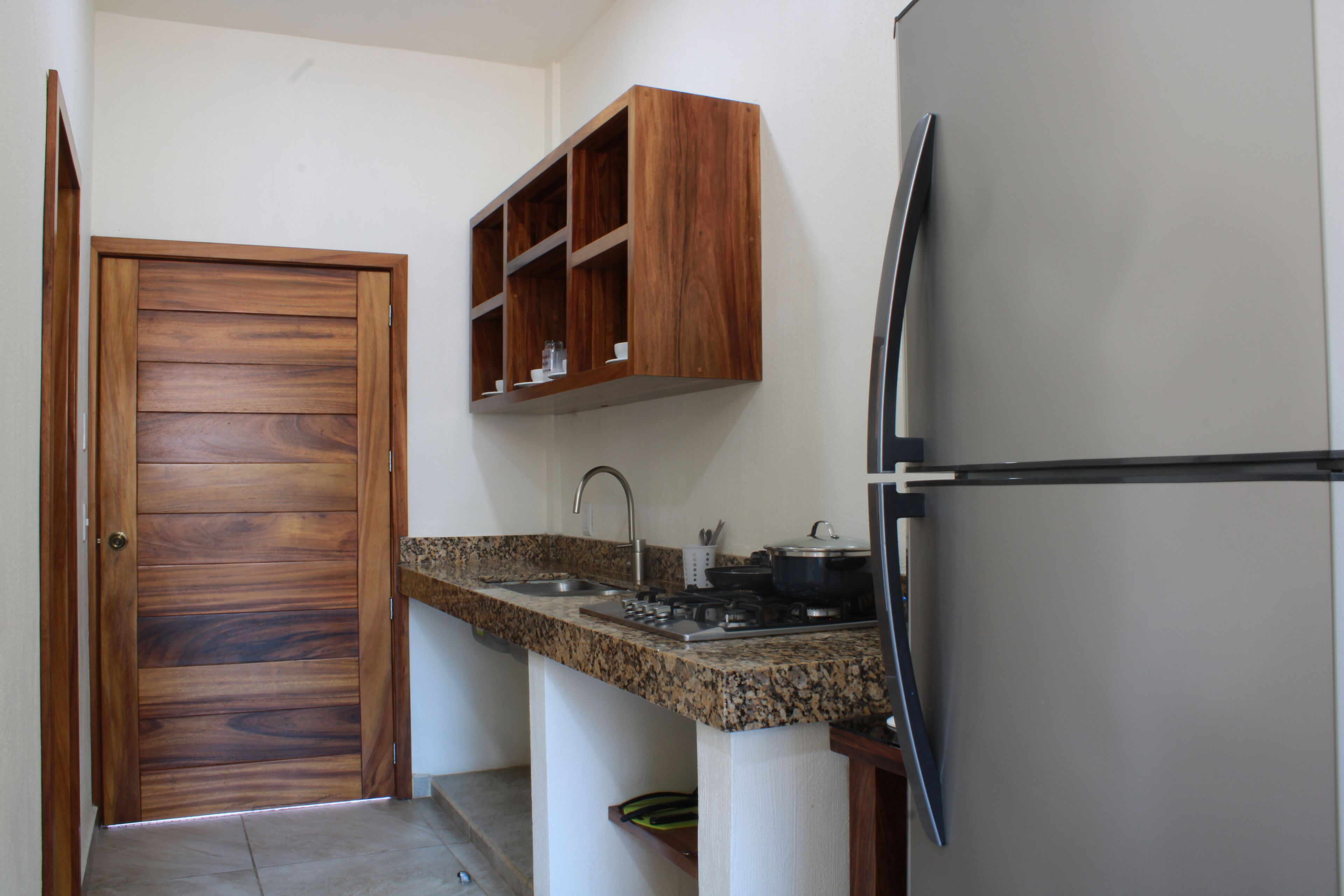 A kitchen with a wooden door, granite countertop, built-in shelves, and a refrigerator.