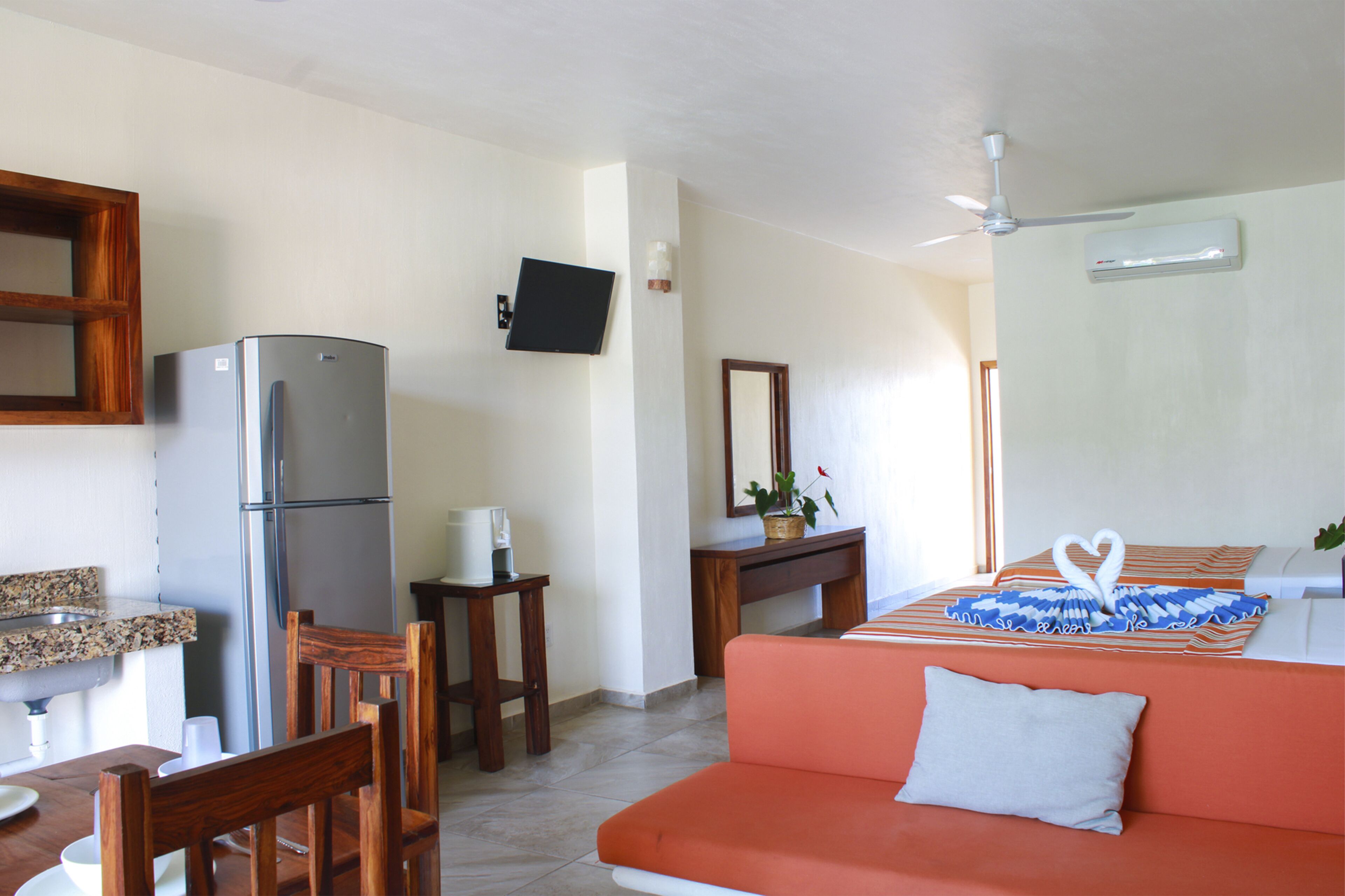 Habitación de hotel compacta con kitchenette, una cama con almohada con forma de corazón, una silla de madera, una mesita pequeña y un televisor montado en la pared.