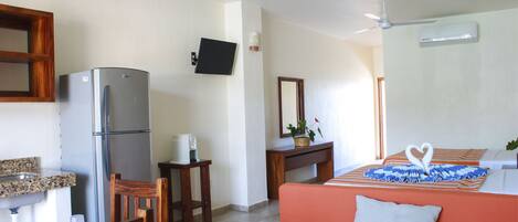 Habitación de hotel compacta con kitchenette, una cama con almohada con forma de corazón, una silla de madera, una mesita pequeña y un televisor montado en la pared.