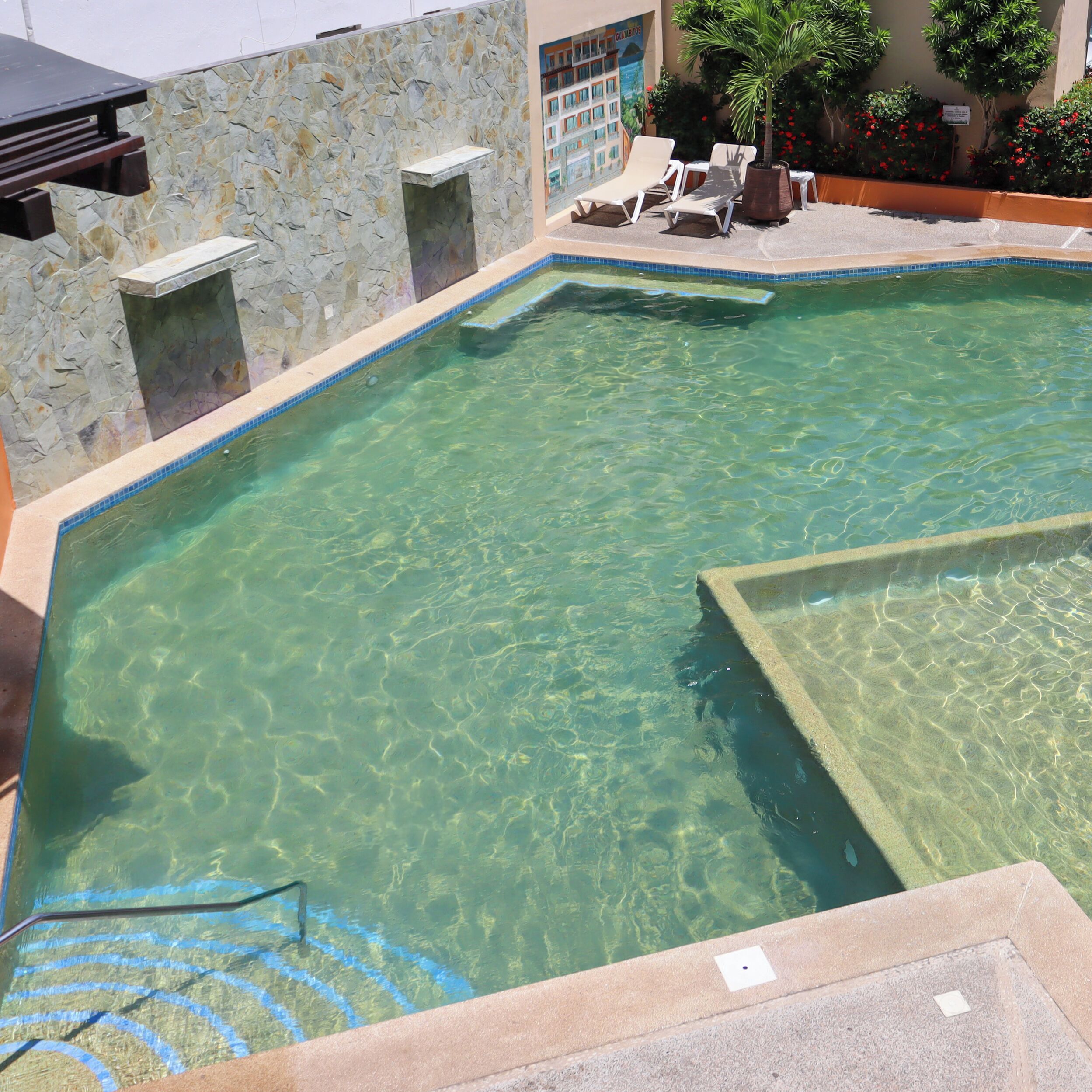 Piscina cristalina con paredes de piedra y zona de descanso con sillas.