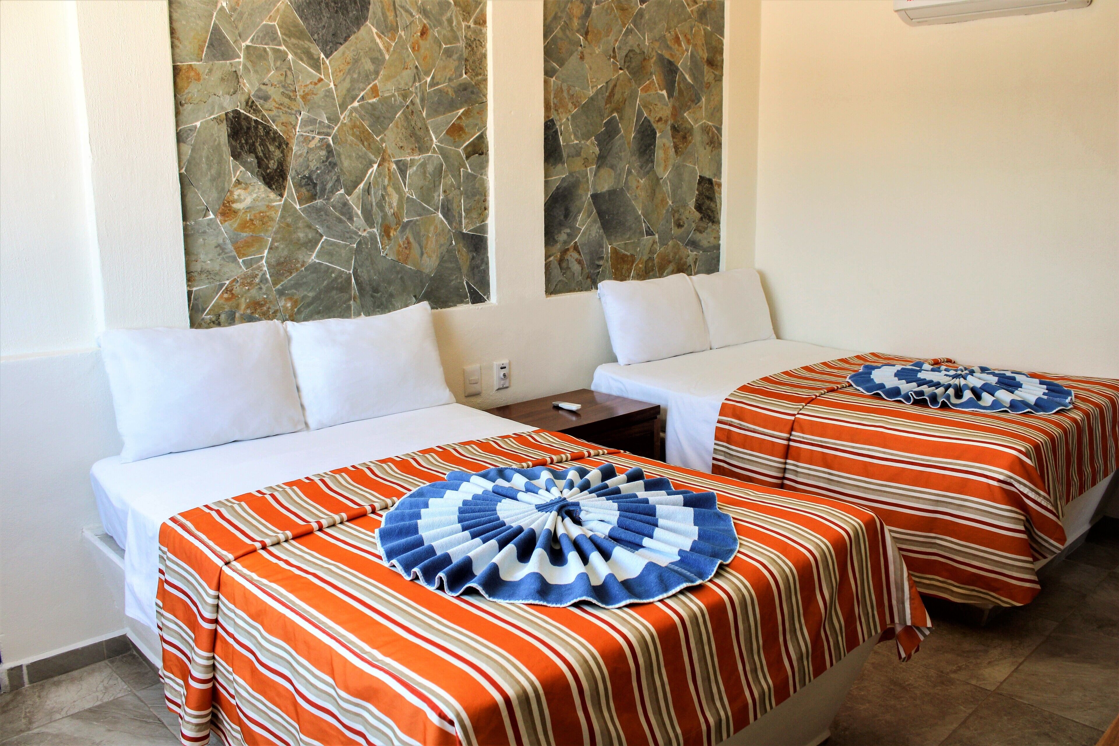 A hotel room with two beds, each with a striped bedspread and a decorative pillow. The room has a stone wall and a small table with a lamp.