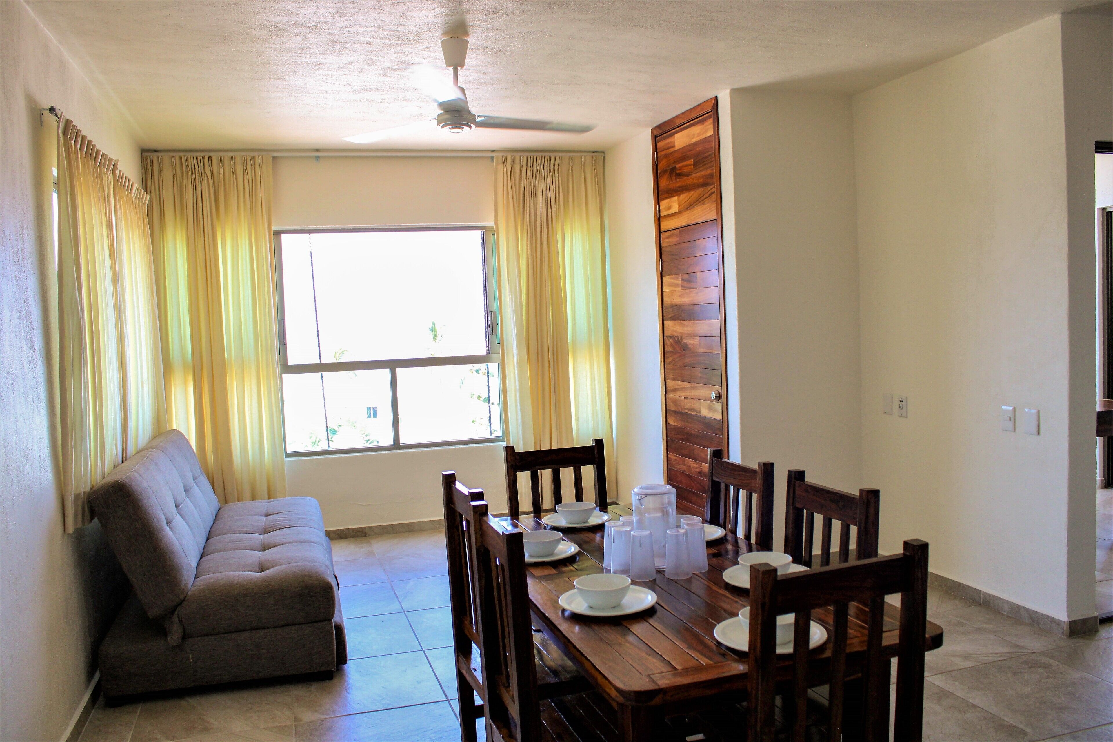 Un comedor con una mesa de madera para cuatro personas, un sofá y un ventanal con cortinas.
