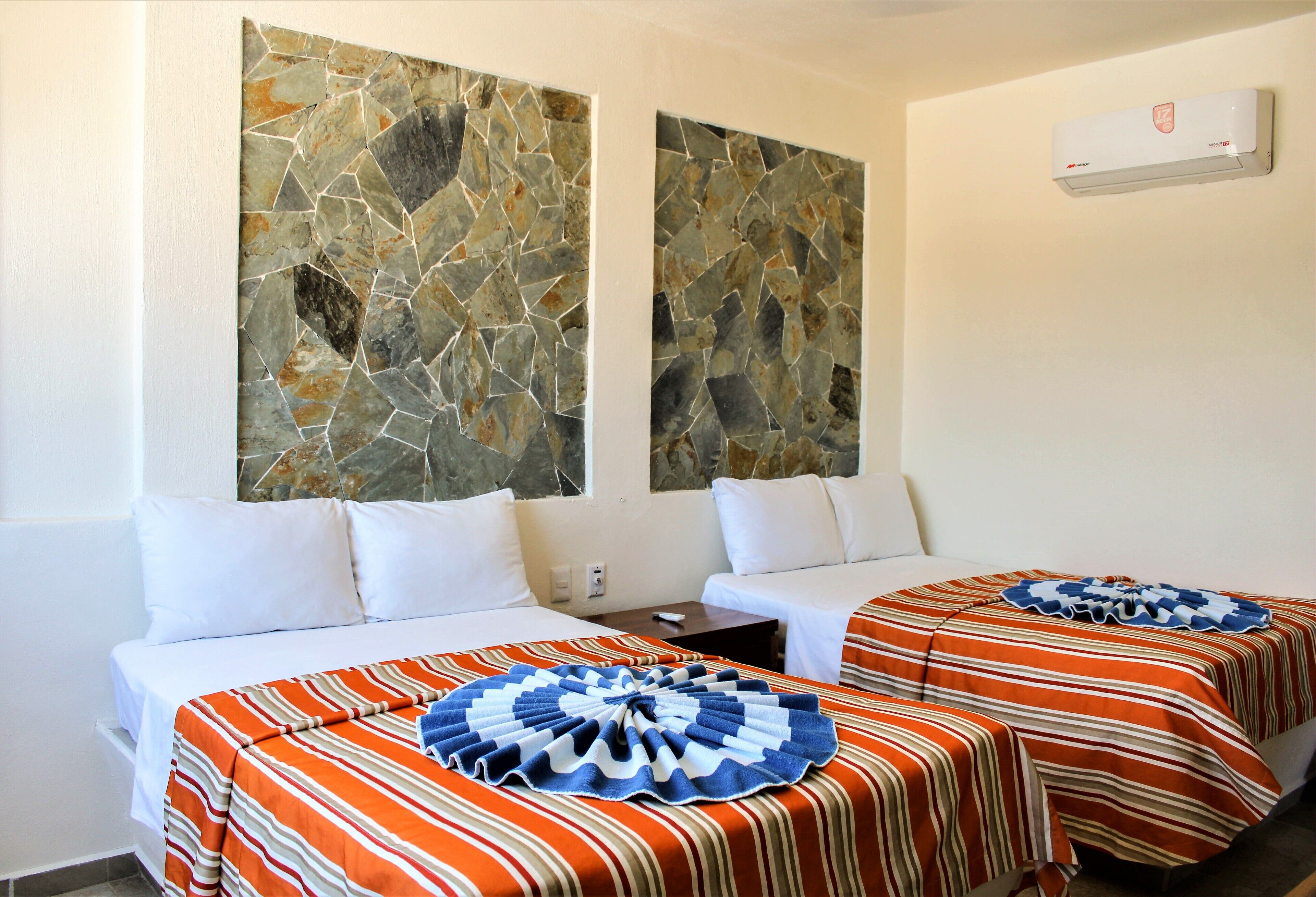 Quarto de hotel com duas camas, cada uma com roupa de cama listrada e almofada decorativa azul e branca. Duas grandes peças de arte em pedra adornam a parede sobre as camas.