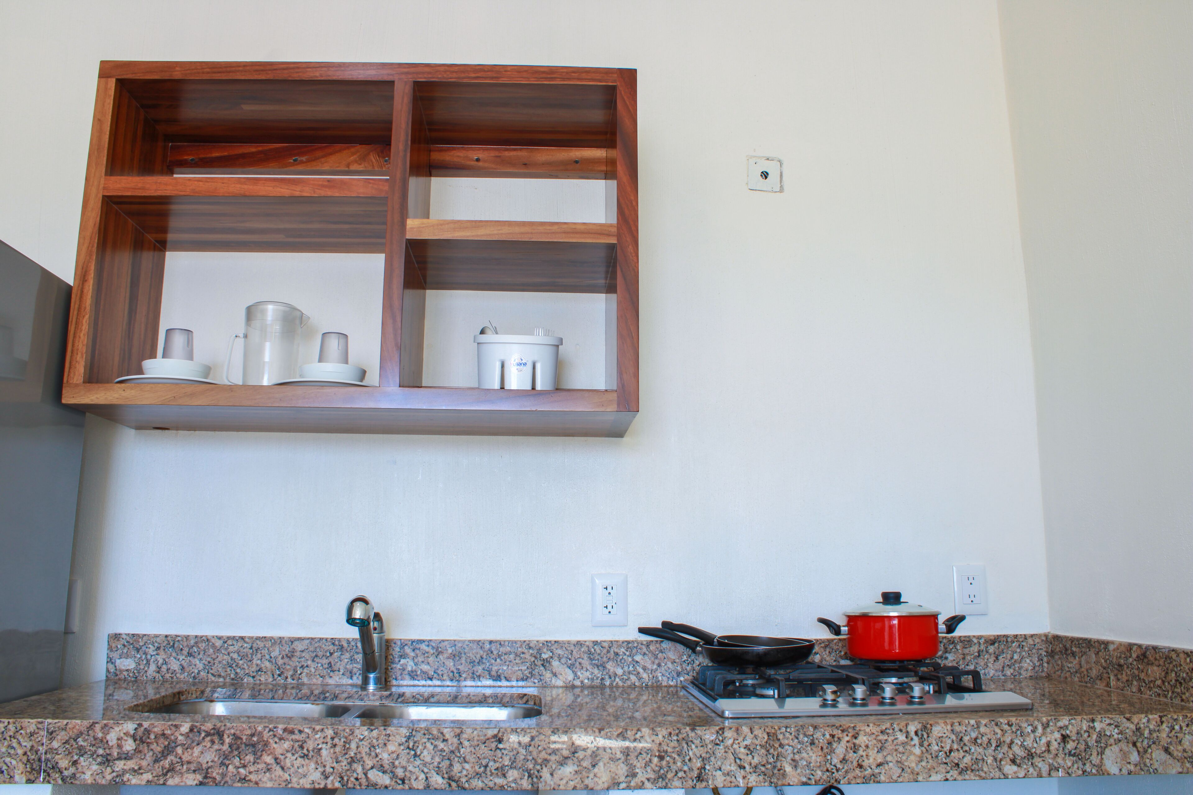 Una cocina con encimeras de granito, un lavamanos de acero inoxidable, una olla roja sobre la estufa y un estante de madera sobre el lavamanos.