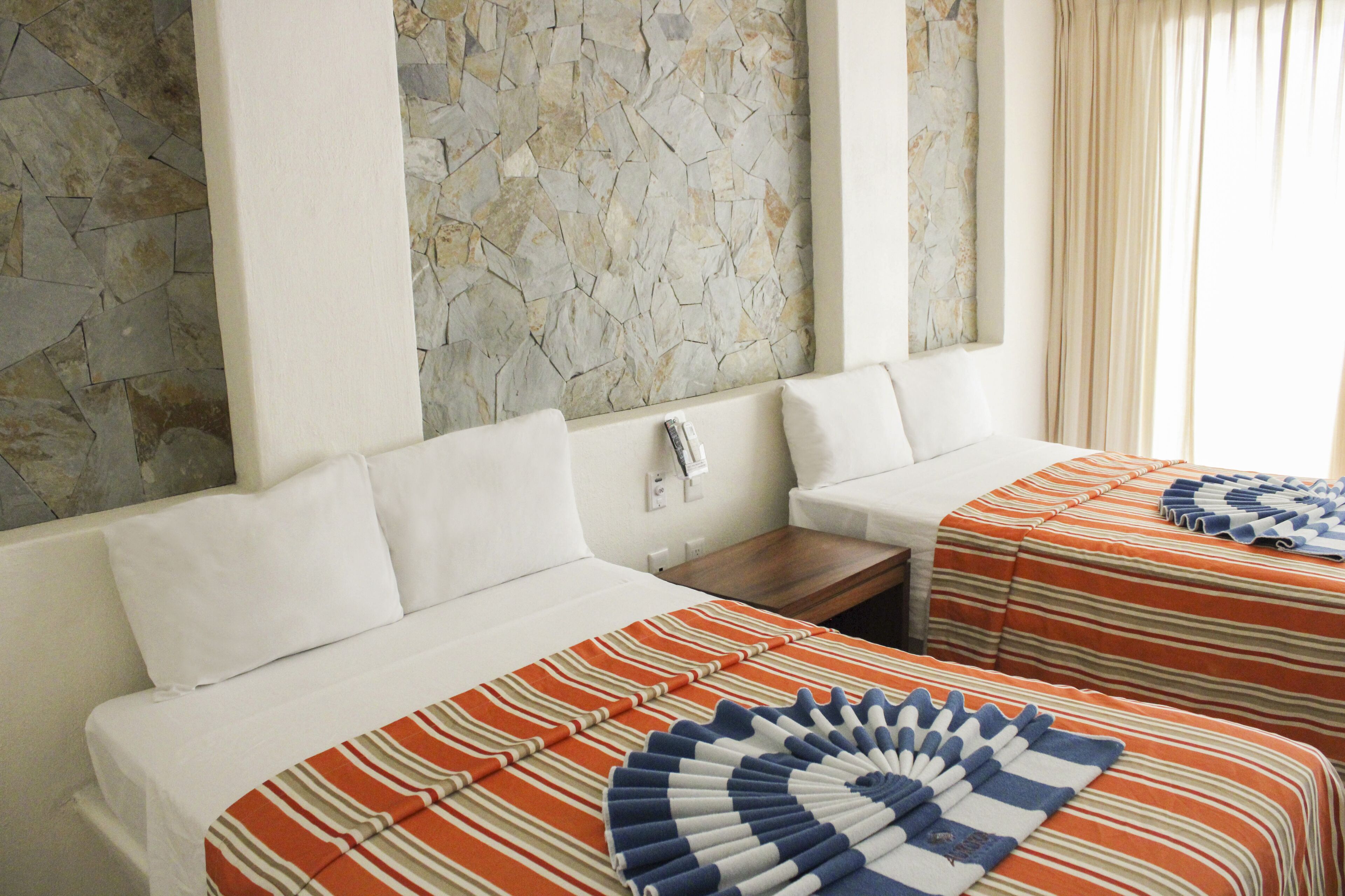 Habitación de hotel con pared de piedra, dos camas con ropa de cama a rayas, almohadas blancas y una mesita de noche de madera.