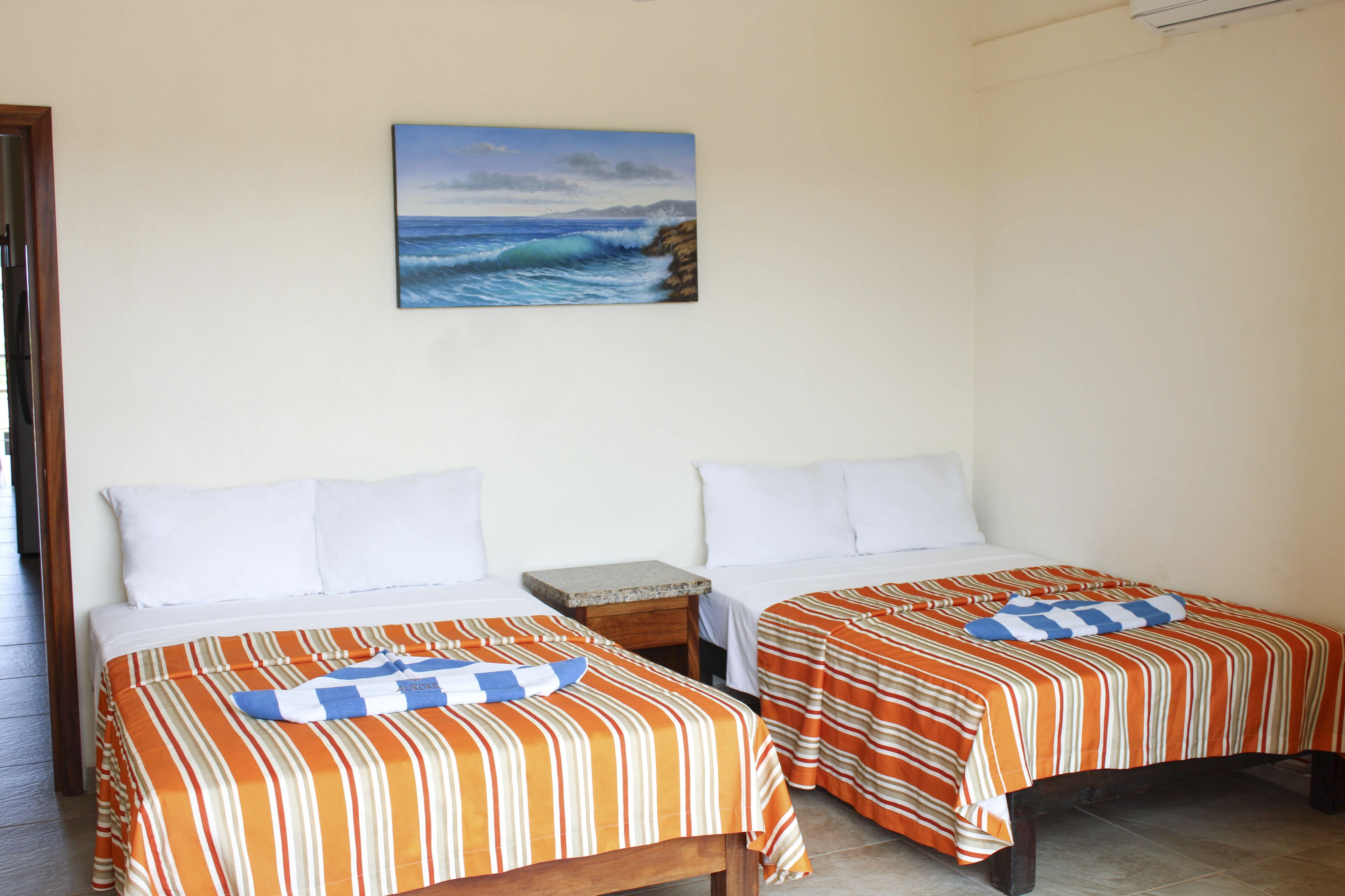 A room with two beds, each with striped bedding, a small bedside table, and a painting of a coastal scene on the wall.