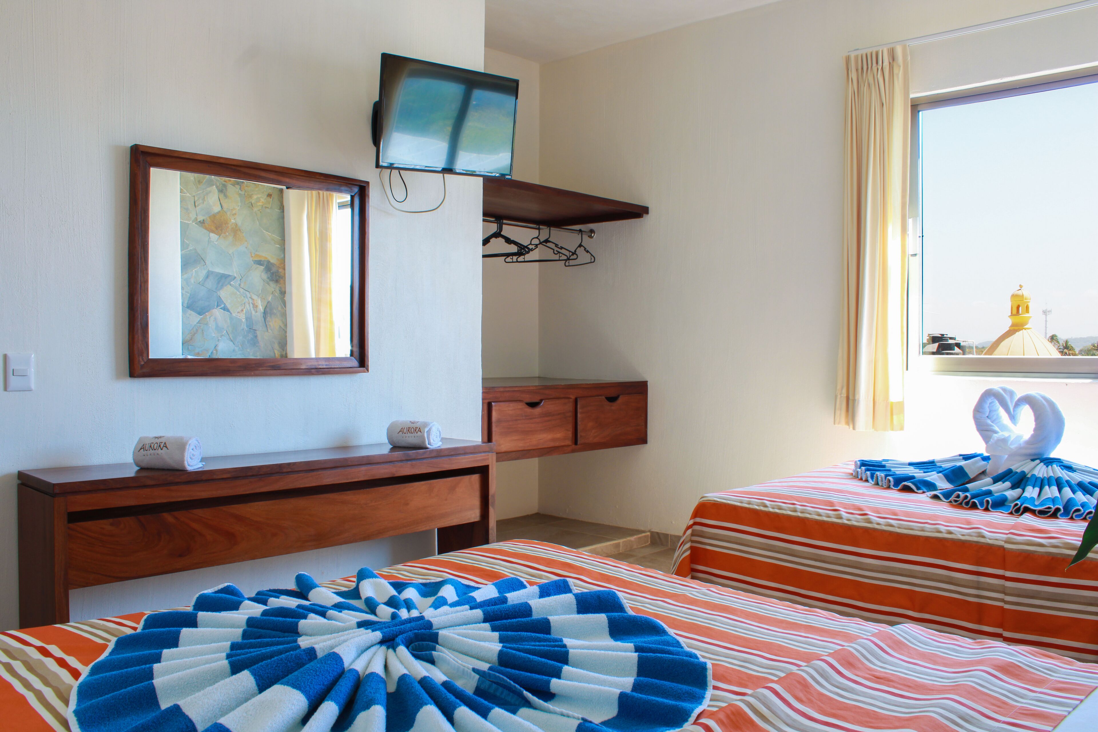 A hotel room with a television mounted on the wall, a wooden dresser, a bed with striped bedding, and a window with curtains.