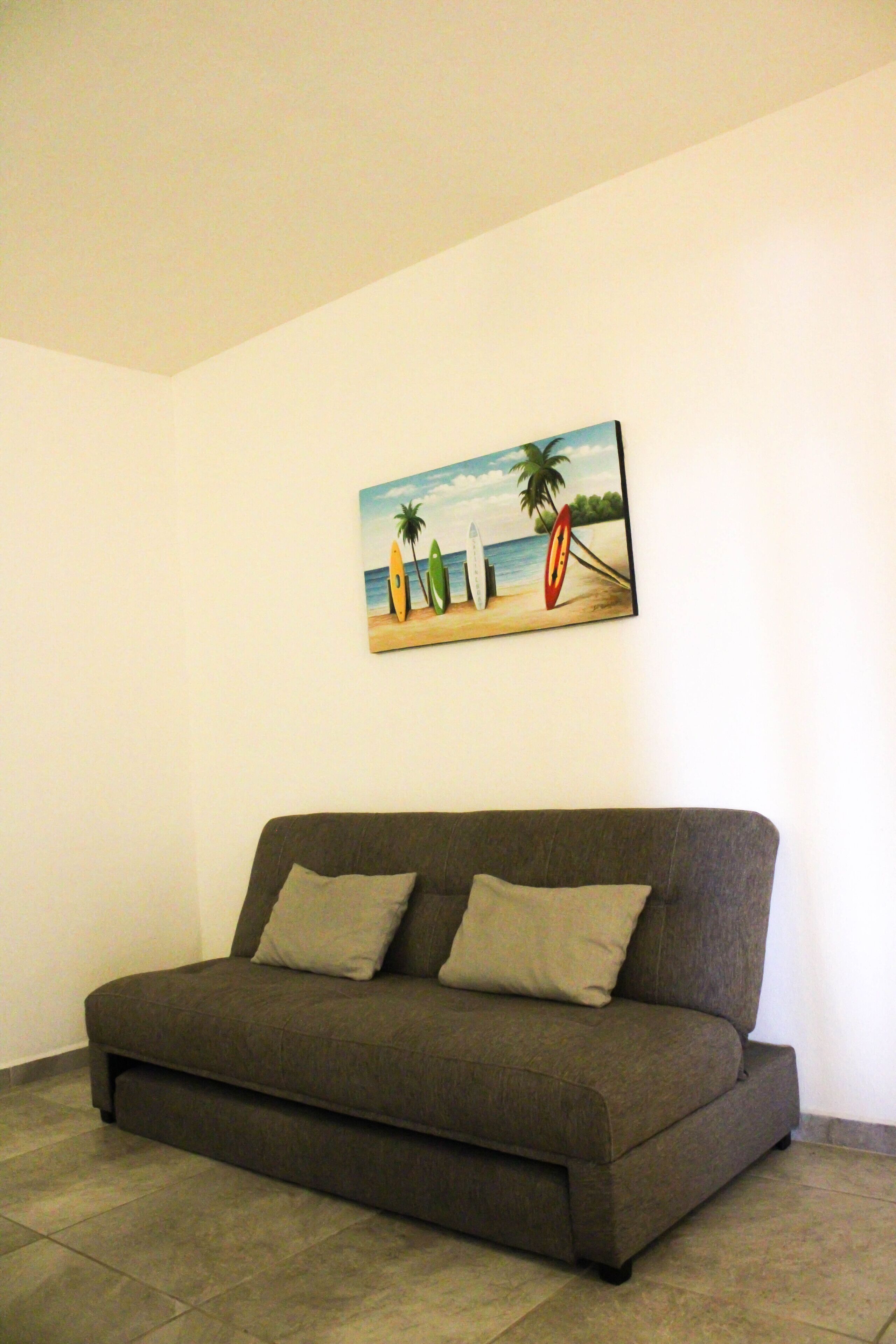Una habitación con un sofá gris, un cuadro de una playa y un suelo de baldosas.
