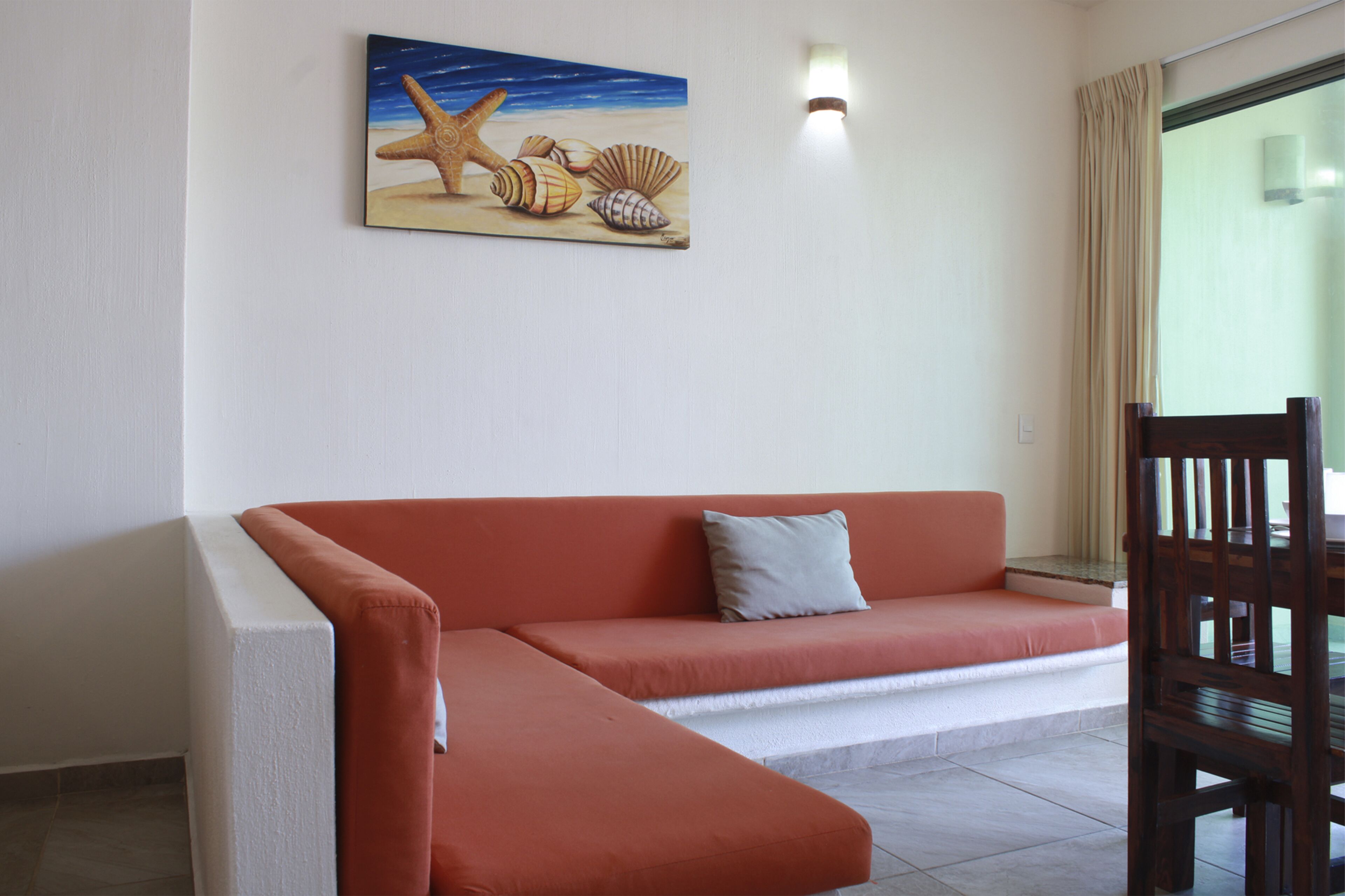 A room with a red sofa, a wooden chair, and a painting of a starfish and seashells on the wall.