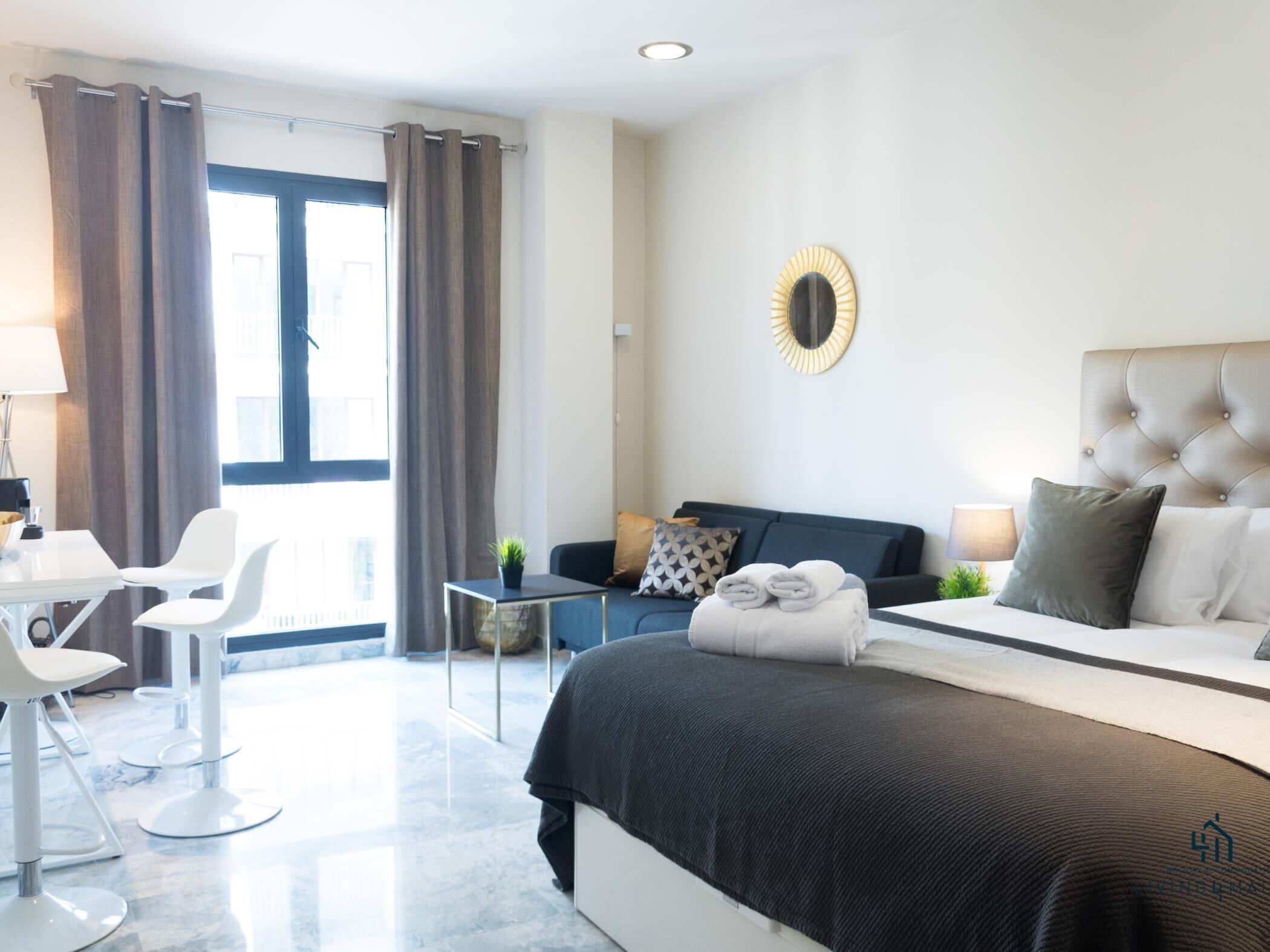 Une chambre d’hôtel moderne équipée d’un lit, d’un canapé, d’un bureau avec des chaises et d’une grande fenêtre avec des rideaux.