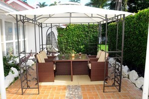 Een terras buiten met een tafel met glazen blad, rieten meubels en een metalen pergola.