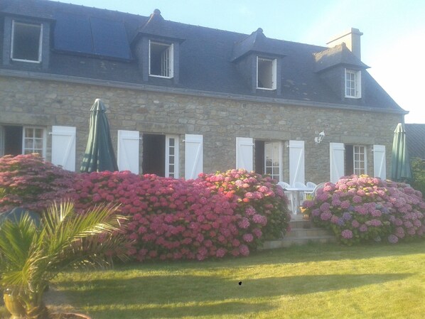 Un bâtiment en pierre doté de nombreuses fenêtres et volets, entouré de haies bien entretenues et d’une pelouse.