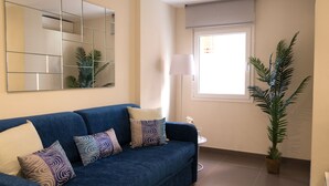 A living room with a blue sofa, patterned cushions, a large mirror, a potted plant, and a window with a view.