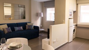 A modern living room with a blue sofa, a dining table with chairs, a flat-screen TV, and a kitchen area with a washing machine and microwave.