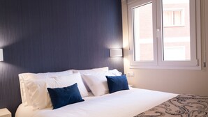 A neatly made bed with white and blue pillows, a patterned throw, and a window with a view of a building.