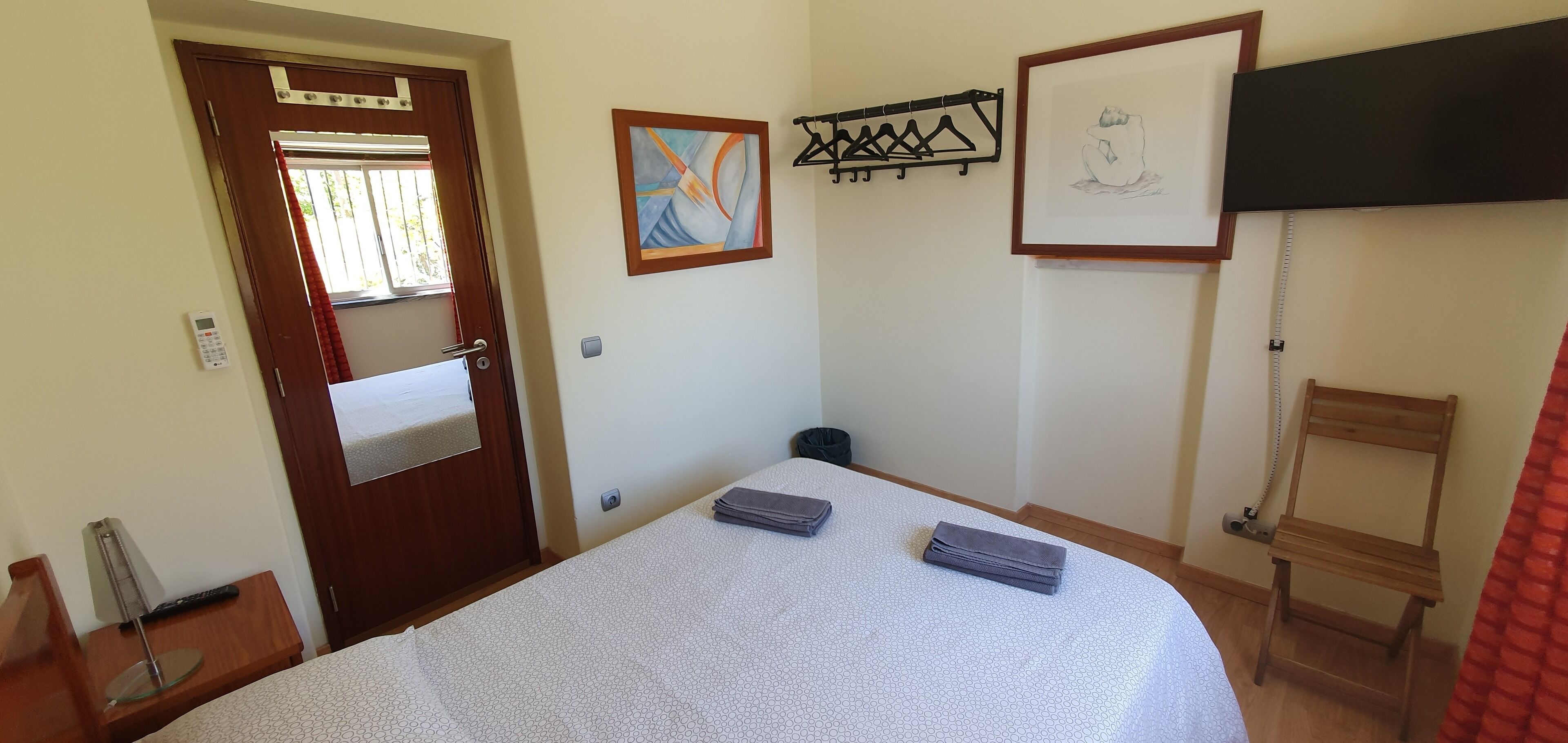 Habitación de hotel con cama, una silla de madera, televisor montado en la pared, un cuadro enmarco y un perchero.