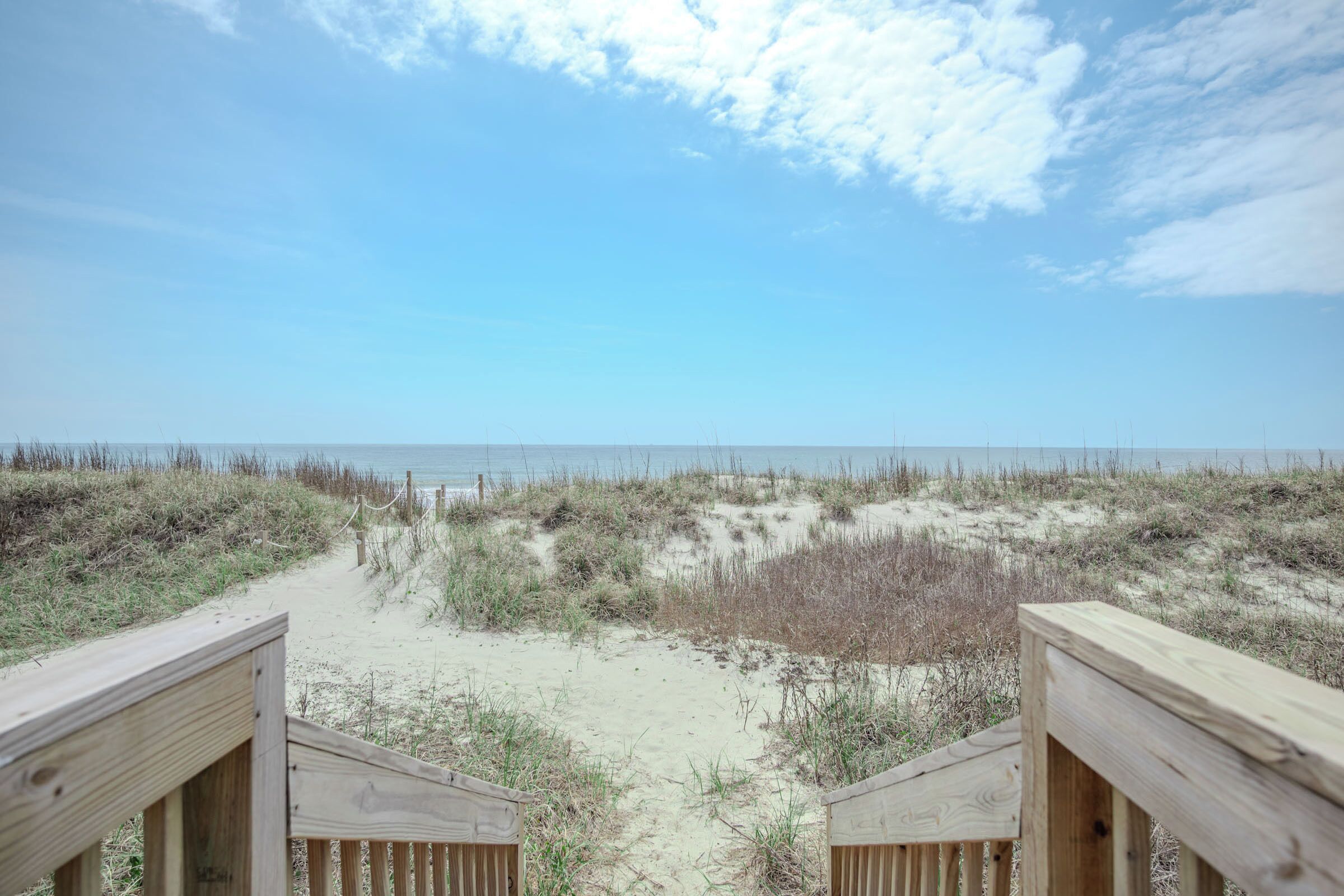Ein hölzerner Steg führt zu einem Strand mit Dünen und spärlicher Vegetation.