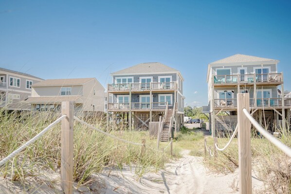 Un bien immobilier situé en bord de mer, composé de plusieurs bâtiments à étages dotés de balcons et d’un chemin de sable menant à l’entrée.