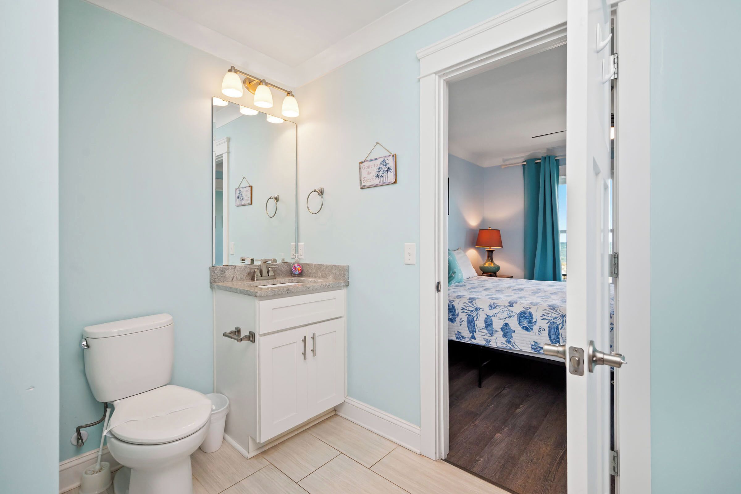 A bathroom with a toilet, sink, and mirror. There is a bed visible through an open door in the adjacent room.