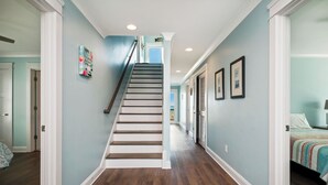 A hallway with a wooden staircase, light blue walls, and a bed with a striped blanket. - Salt & Light: Spacious, Custom-built, Oceanfront Home Close to Splash Pad, Park (Oak Island)