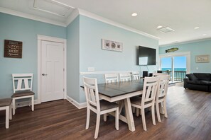 Un coin repas avec une table et des chaises, une porte blanche, un téléviseur fixé au mur et une vue sur l’océan par une porte vitrée.