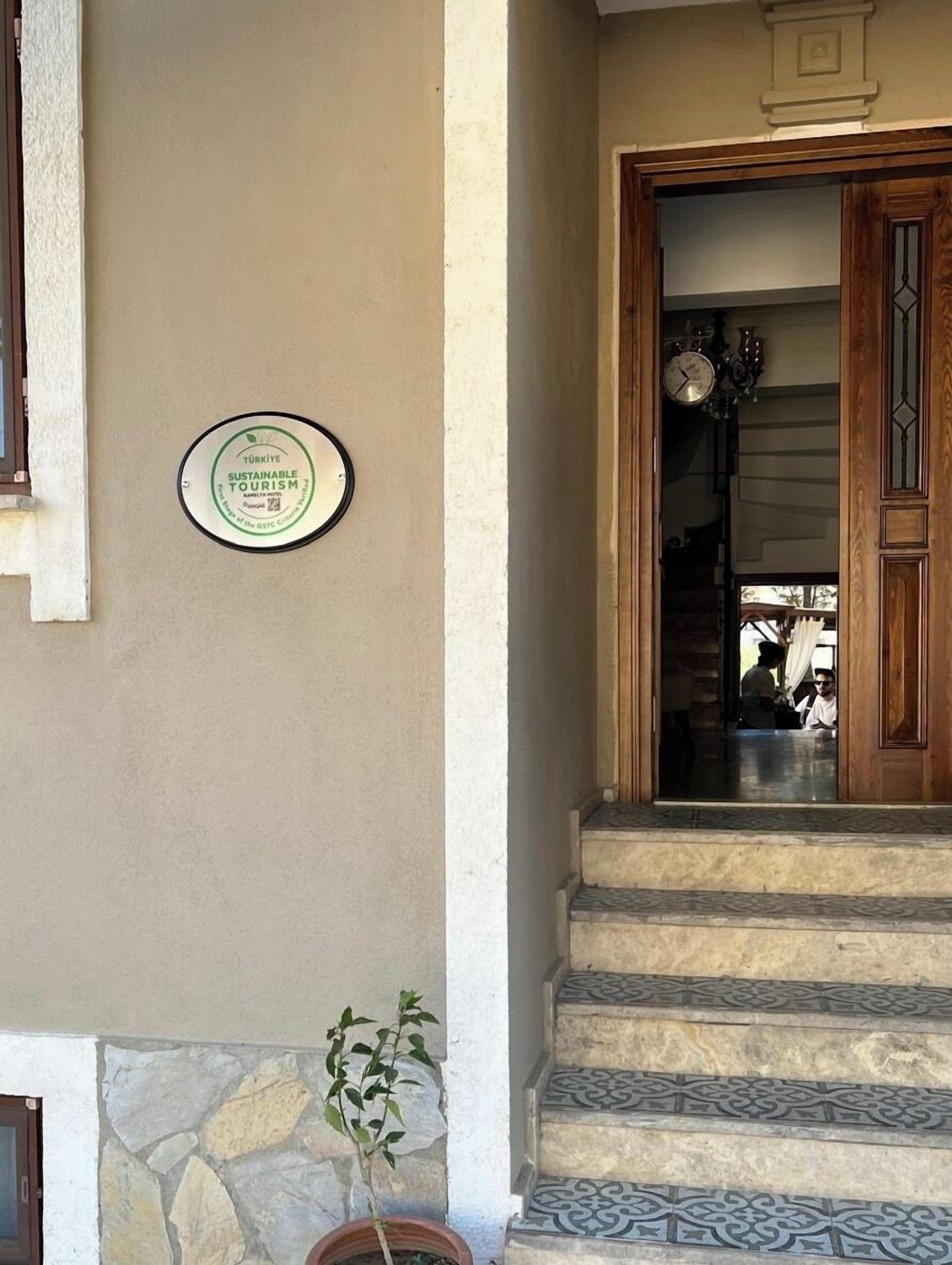 A building entrance with a sign indicating sustainable tourism, a stone wall, a potted plant, and a staircase with patterned tiles.