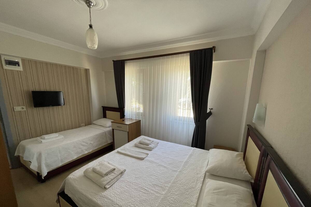 A hotel room with two single beds, a flat-screen TV mounted on the wall, a bedside table with a lamp, and a window with sheer curtains.