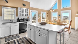 A modern kitchen with white cabinets, a central island, and a dining area with wooden chairs. - Sandtrap: Oceanview House on West Beach with Easy Beach Access. (Oak Island)