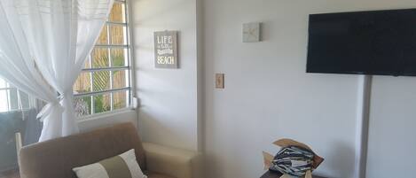 A living room with a beige sofa, a flat-screen TV mounted on the wall, and a small wooden table with decorative items.