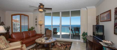 A living room with a sofa, coffee table, and TV, a balcony with a view of the ocean, and a ceiling fan.