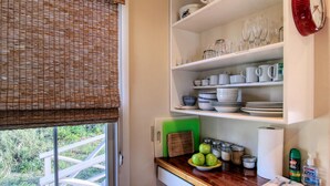Una cocina con ventana, repisas con vajilla, una tabla de cortar y un bol con manzanas.
